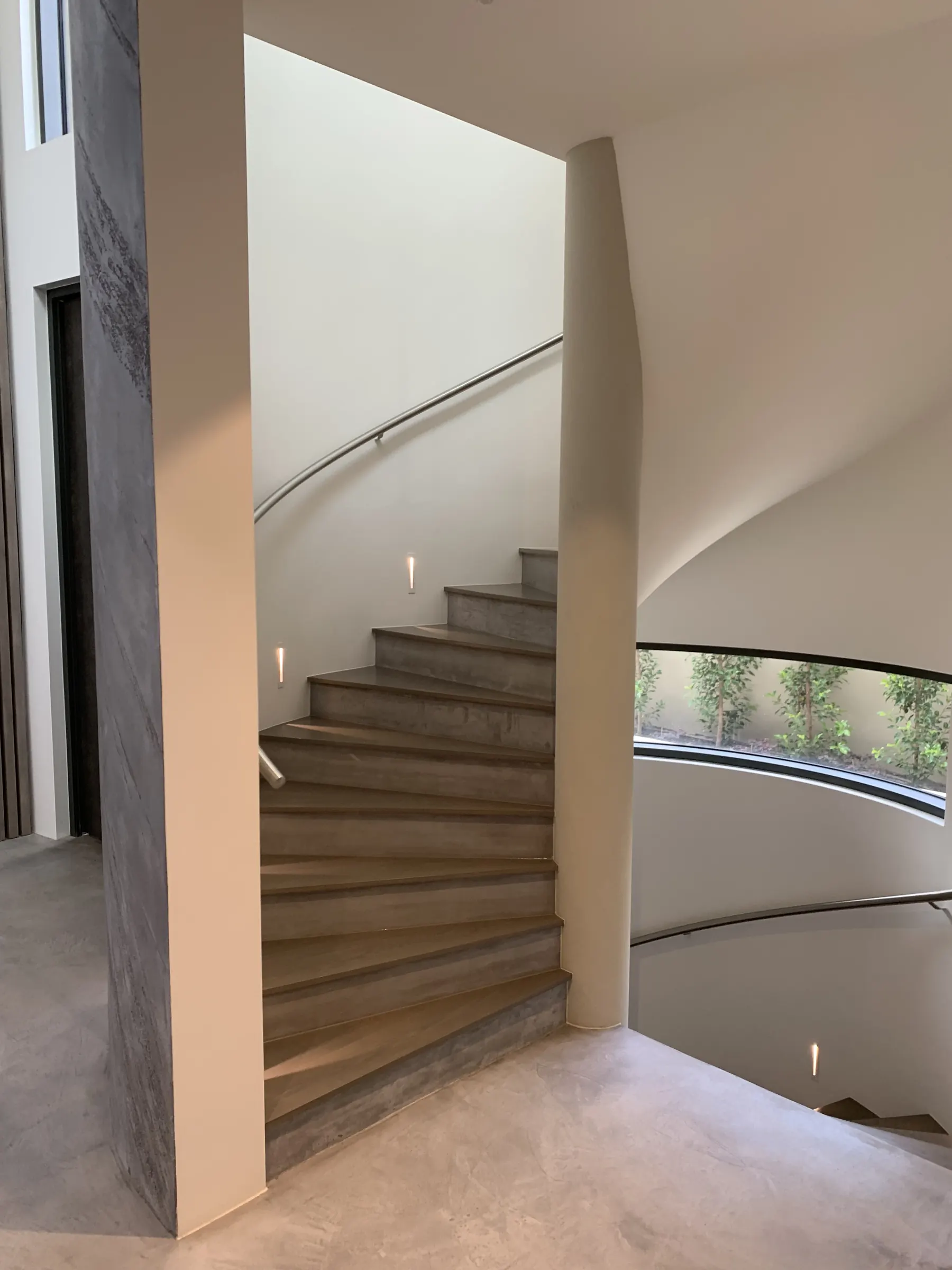 Curved concrete stair with a steel handrail and a window onto the garden