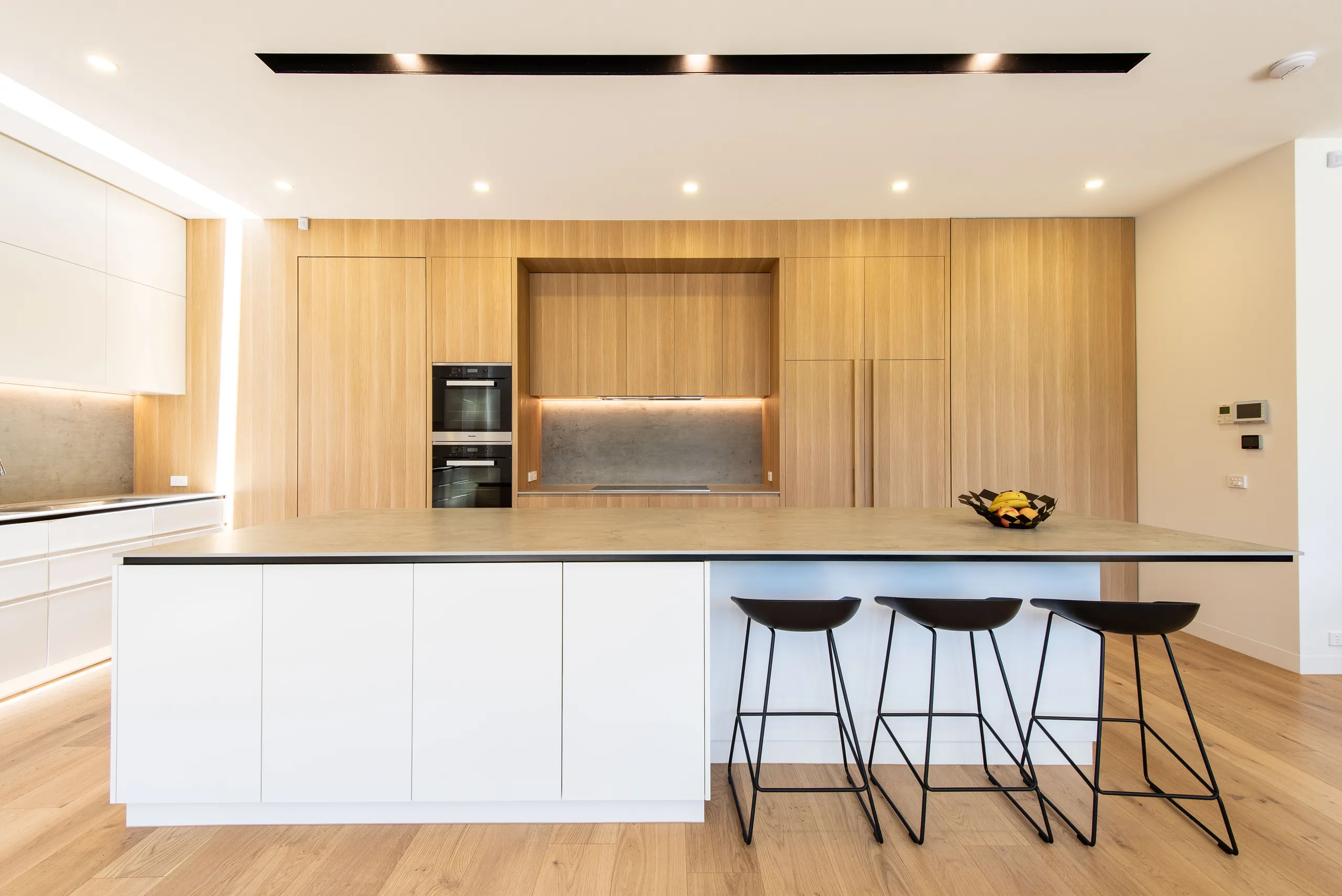 Kitchen island in white and stone with stools beneath a timber joinery wall